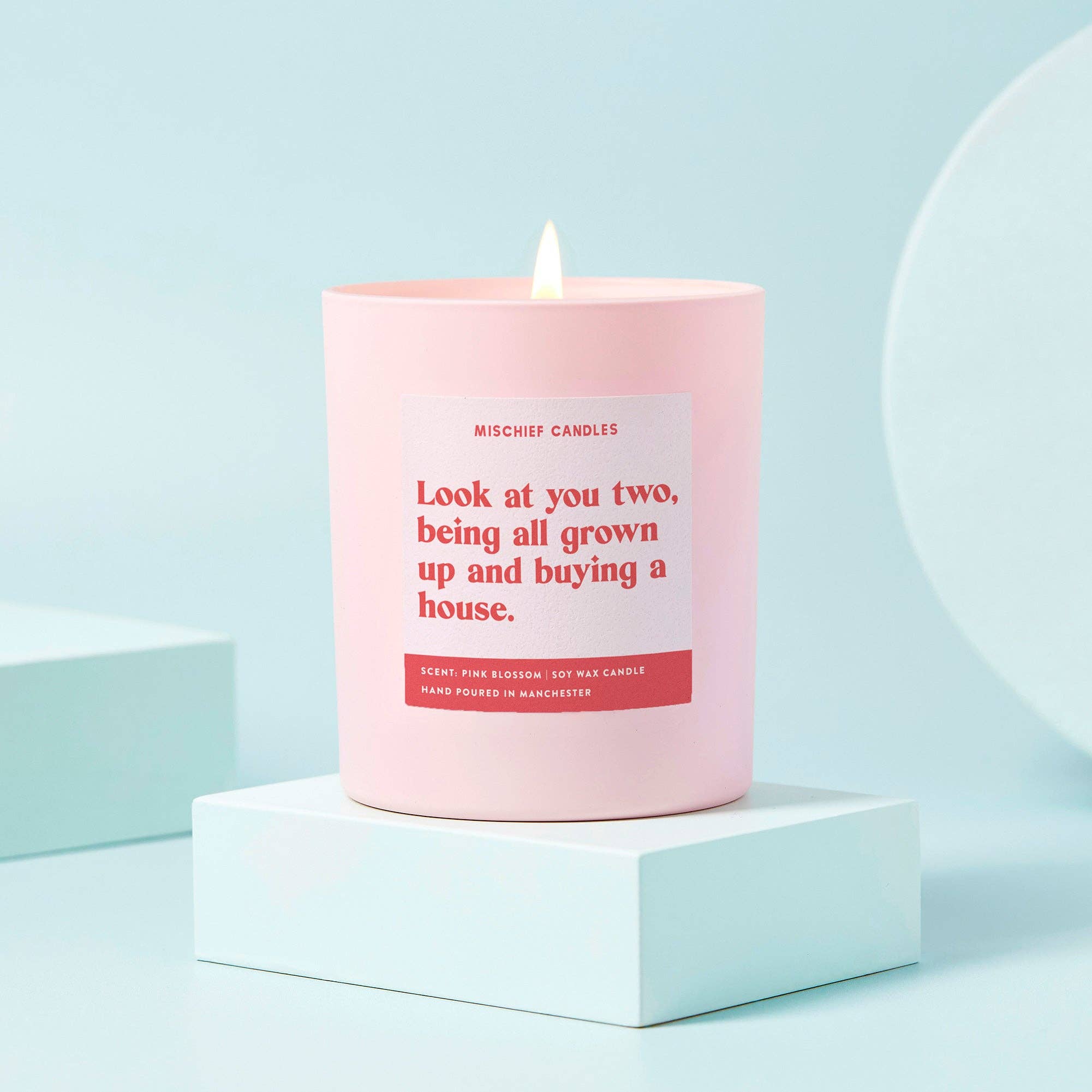 A pale pink candle with red text label reading 'Look at you two, being all grown up and buying a house.' with a lit flame, placed on a white surface.