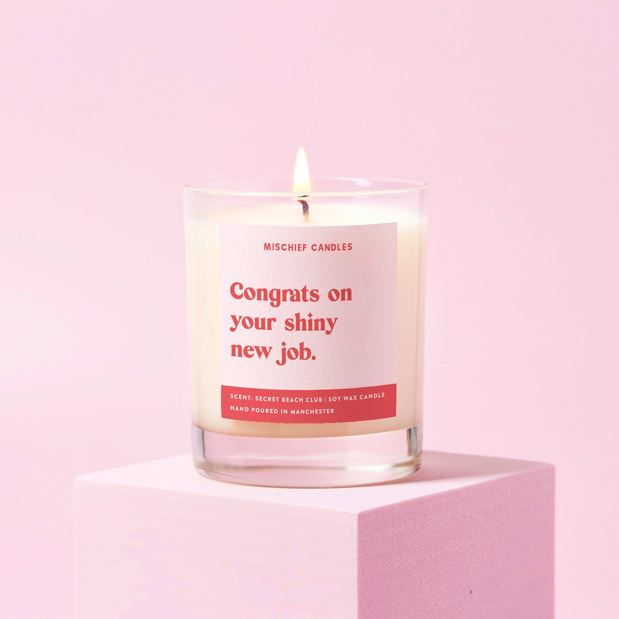 A pale pink candle with the text 'Congrats on your shiny new job' written in bold red font on the label. The candle is lit and placed on a pink surface with a white background.